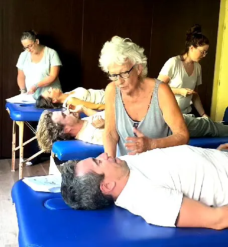 Atelier pratique : une formatrice pose les mains sur un participant allongé sur une table de massage, d’autres binômes travaillent derrière